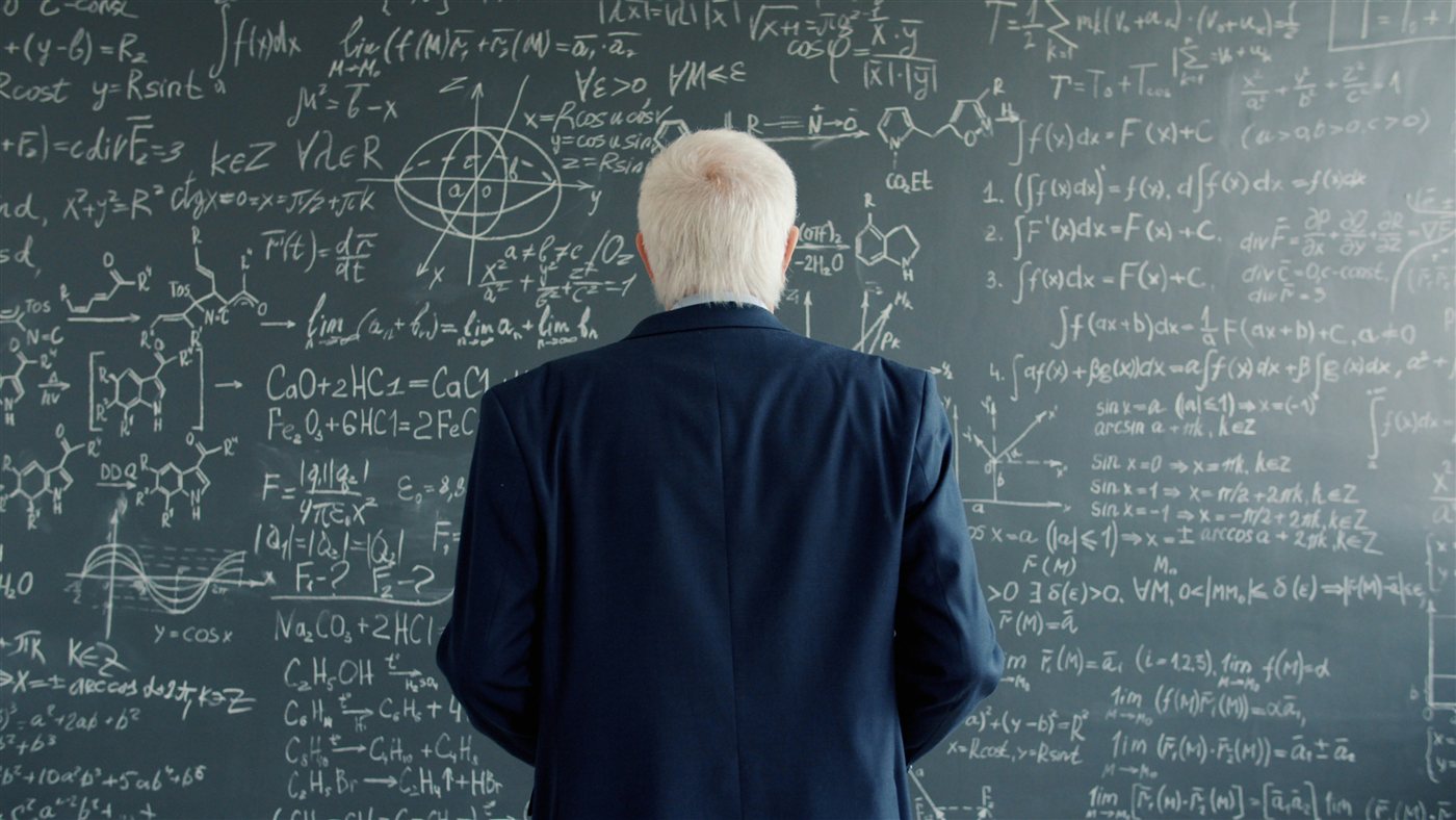 A professor working through physics equations on a blackboard