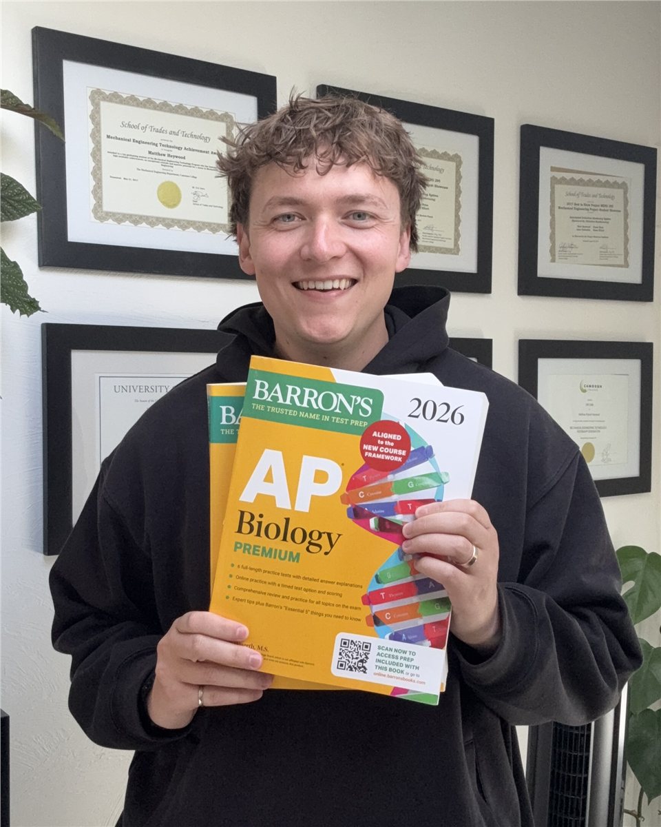 Matt Heywood holding AP Biology prep books, with Camosun College and University of Victoria diplomas on the wall behind him